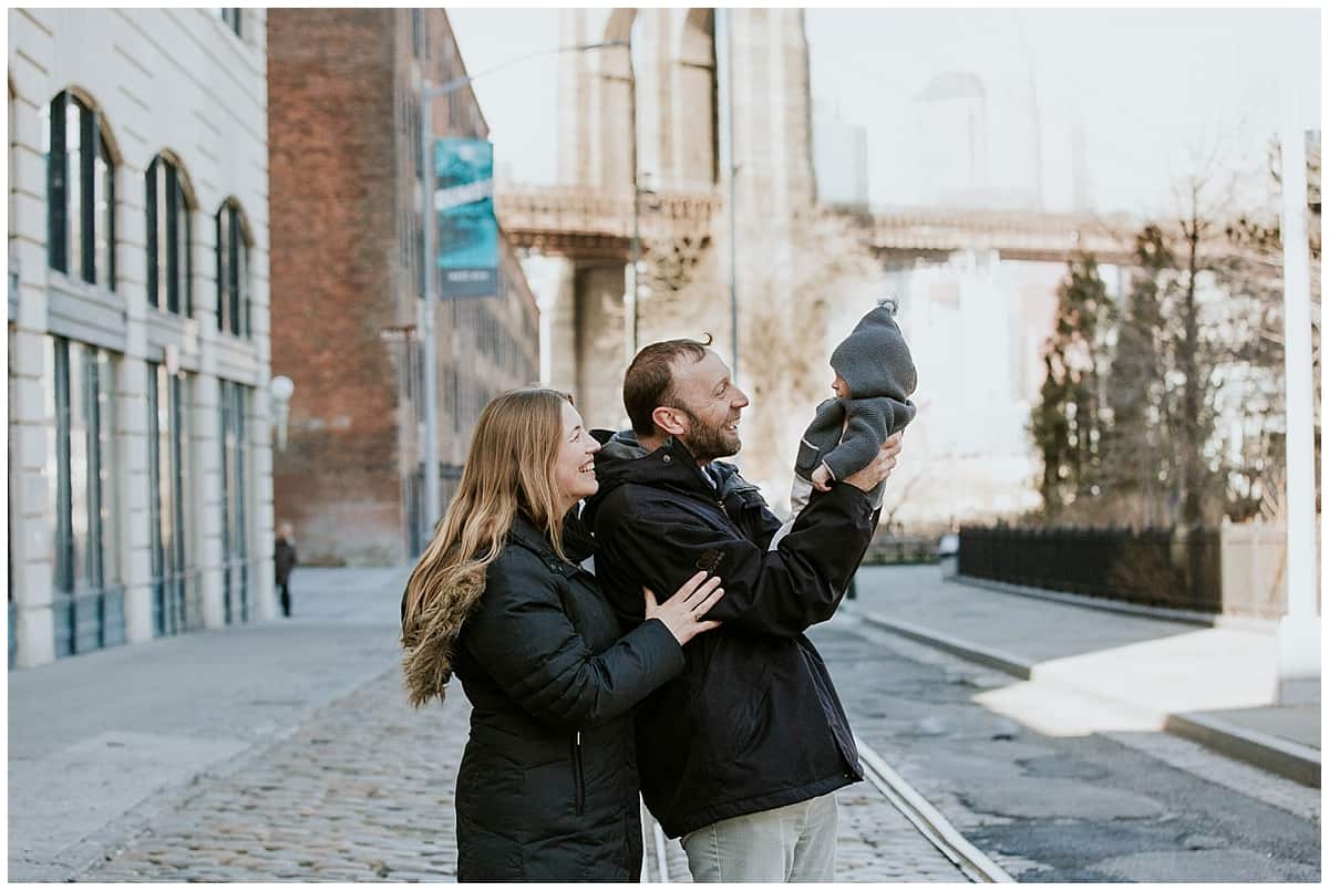 One of the best places to visit with a baby is New York - parents holding a baby in front of the Brooklyn Bridge