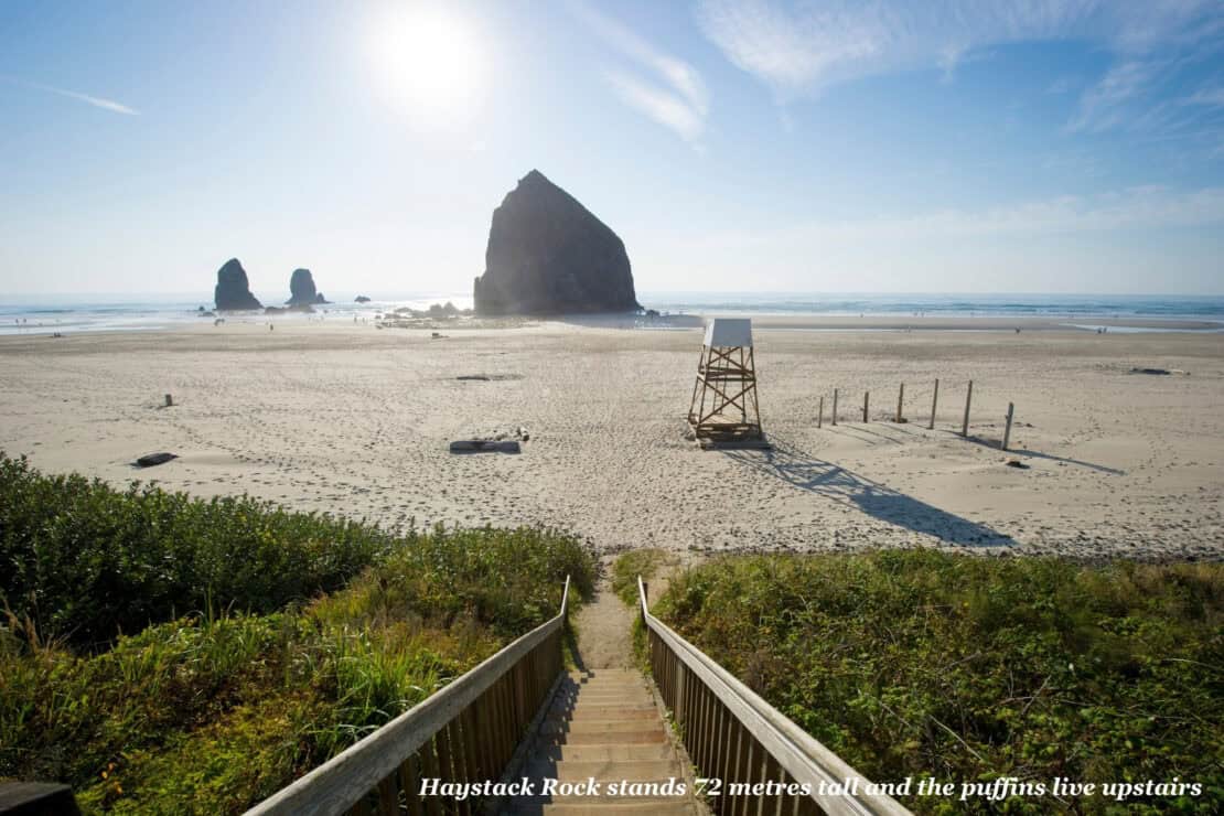 Wooden steps leading down through dune grass to the wide sand of Cannon Beach, Oregon, with Haystack Rock and a lifeguard tower in the sun
