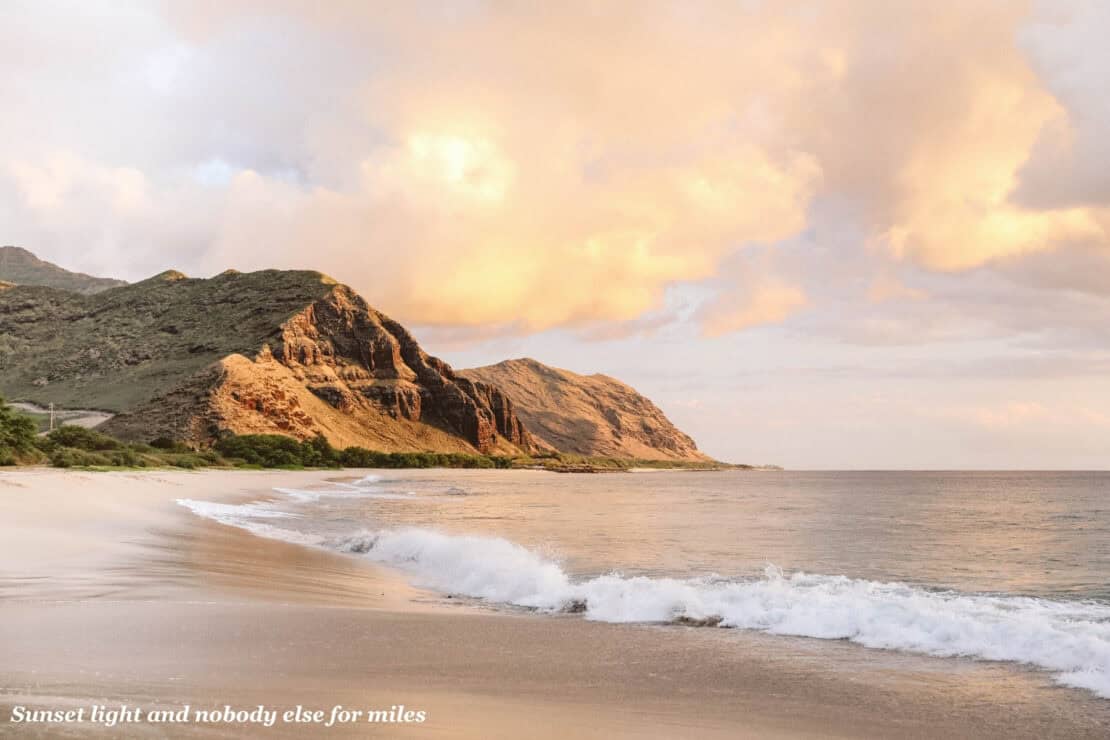 A wide sandy beach on Oahu, Hawaii, with waves rolling in beneath a rugged ridge lit gold by the evening sun.