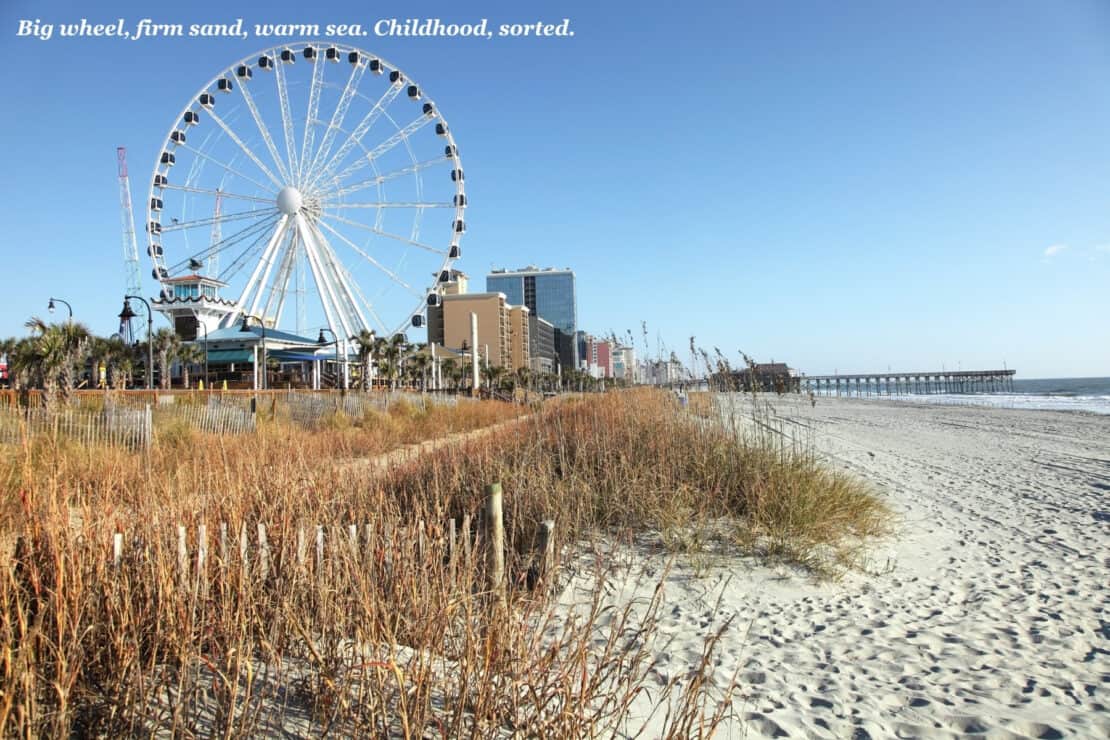 SkyWheel and beachfront hotels behind dune grass and wide sand at Myrtle Beach, South Carolina, with a fishing pier in the distance