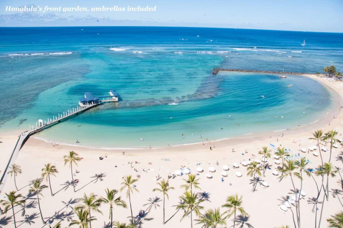 Aerial view of Waikiki Beach in Honolulu, with palm trees, beach umbrellas and a calm lagoon protected by a breakwater